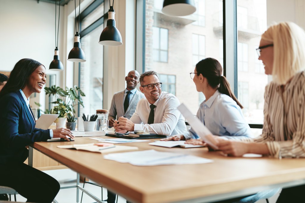 Diverse group of businesspeople laughing while discussing paperwork together during a meeting around a table in a modern office