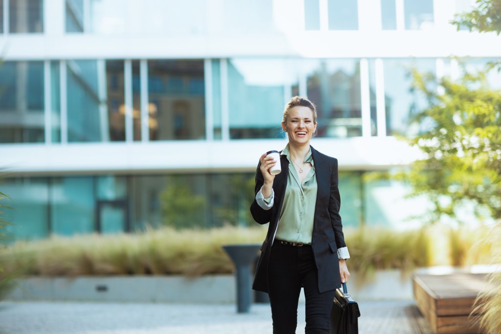 happy modern woman worker in business district in black jacket with cup of coffee walking.