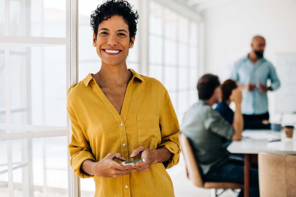 Business woman smiling at the camera and holding a cellphone. Professional business woman standing in an office. Colleagues having a boardroom meeting in the background.