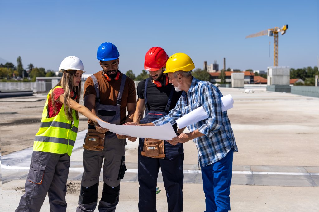 Senior Construction Engineer is Holding a Paper Plans and Doing Analysis with his Younger Coworkers.